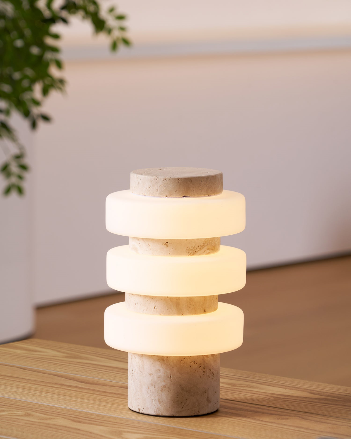 The Clérachic table lamp lit warm on a wooden table, with a green plant at the left and a pale wall behind.