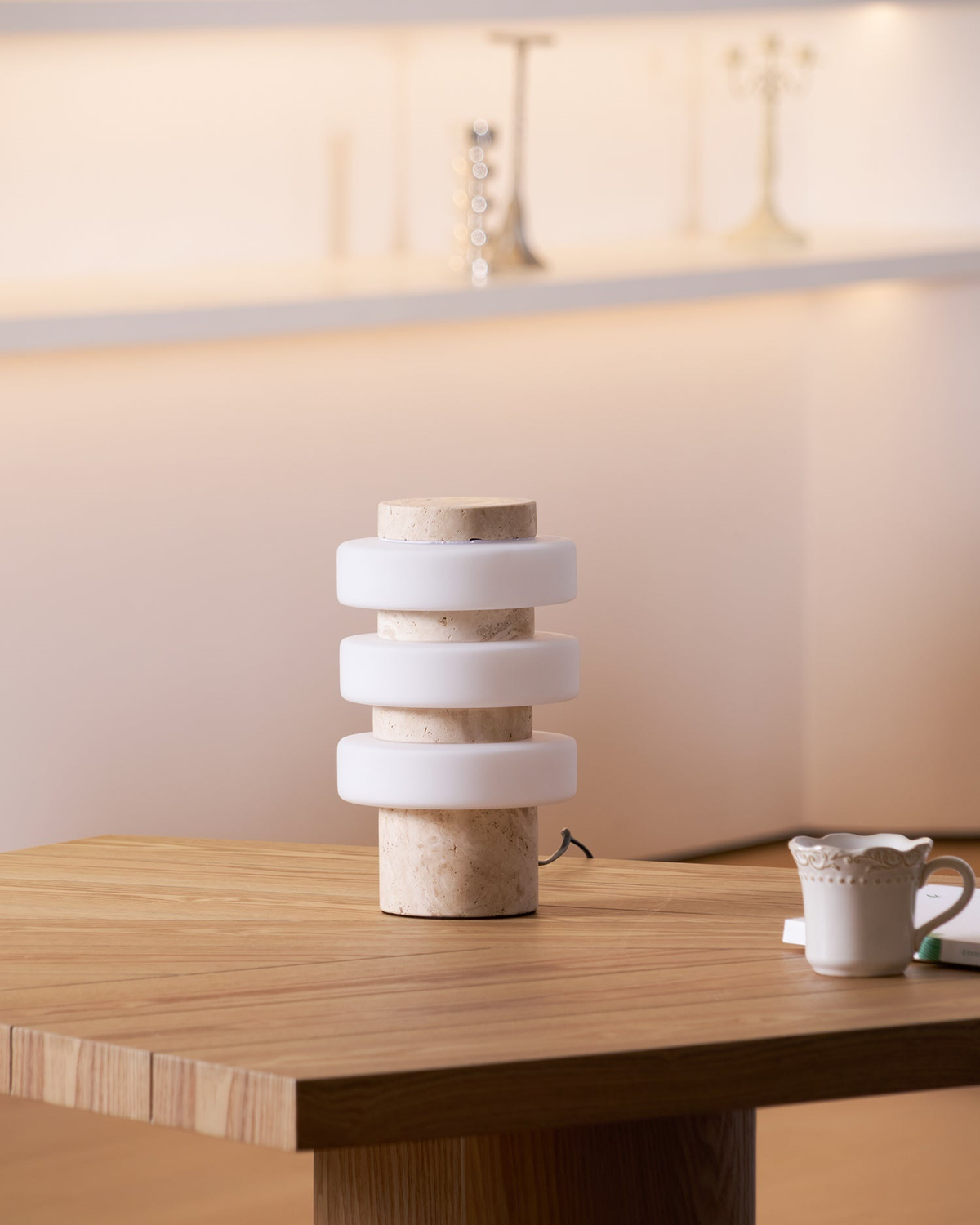 The unlit Clérachic table lamp on a wooden table beside a white china cup, its white glass discs and beige stone base clearly visible.