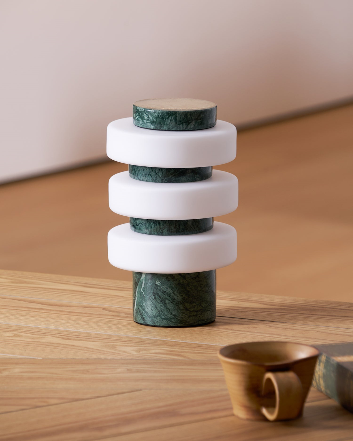 An unlit Clérachic table lamp in the green and white finish on a wooden table, with a brown ceramic cup in the foreground.