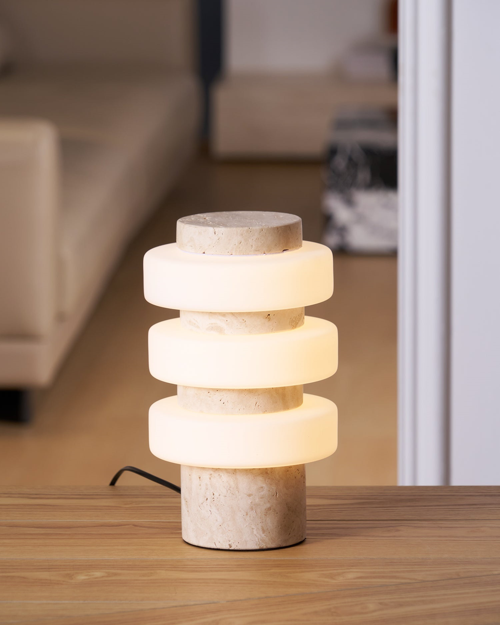The lit Clérachic table lamp on a wooden table in front of a cream sofa, its beige stone base and black cord just visible.