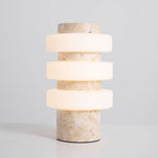 Front view of the lit Clérachic table lamp on a pale grey backdrop, three glowing glass discs on a veined beige stone column.