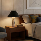 Lit Clérachic marble table lamp on a walnut bedside cabinet, glowing beside a bed with mustard and navy cushions.