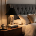 Soft light from a Clérachic marble table lamp on a nightstand, next to a stack of books and a small vase, with a tufted grey headboard behind.