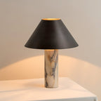 Lit Clérachic table lamp on a white table, the inside of its black shade glowing gold and warming the wall behind.