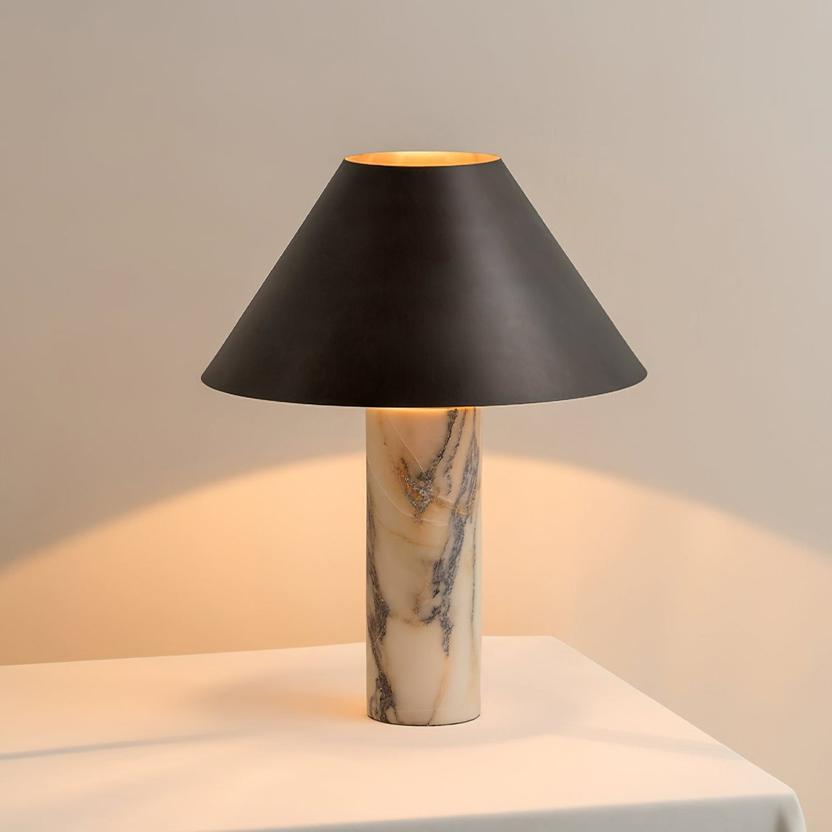 Lit Clérachic table lamp on a white table, the inside of its black shade glowing gold and warming the wall behind.
