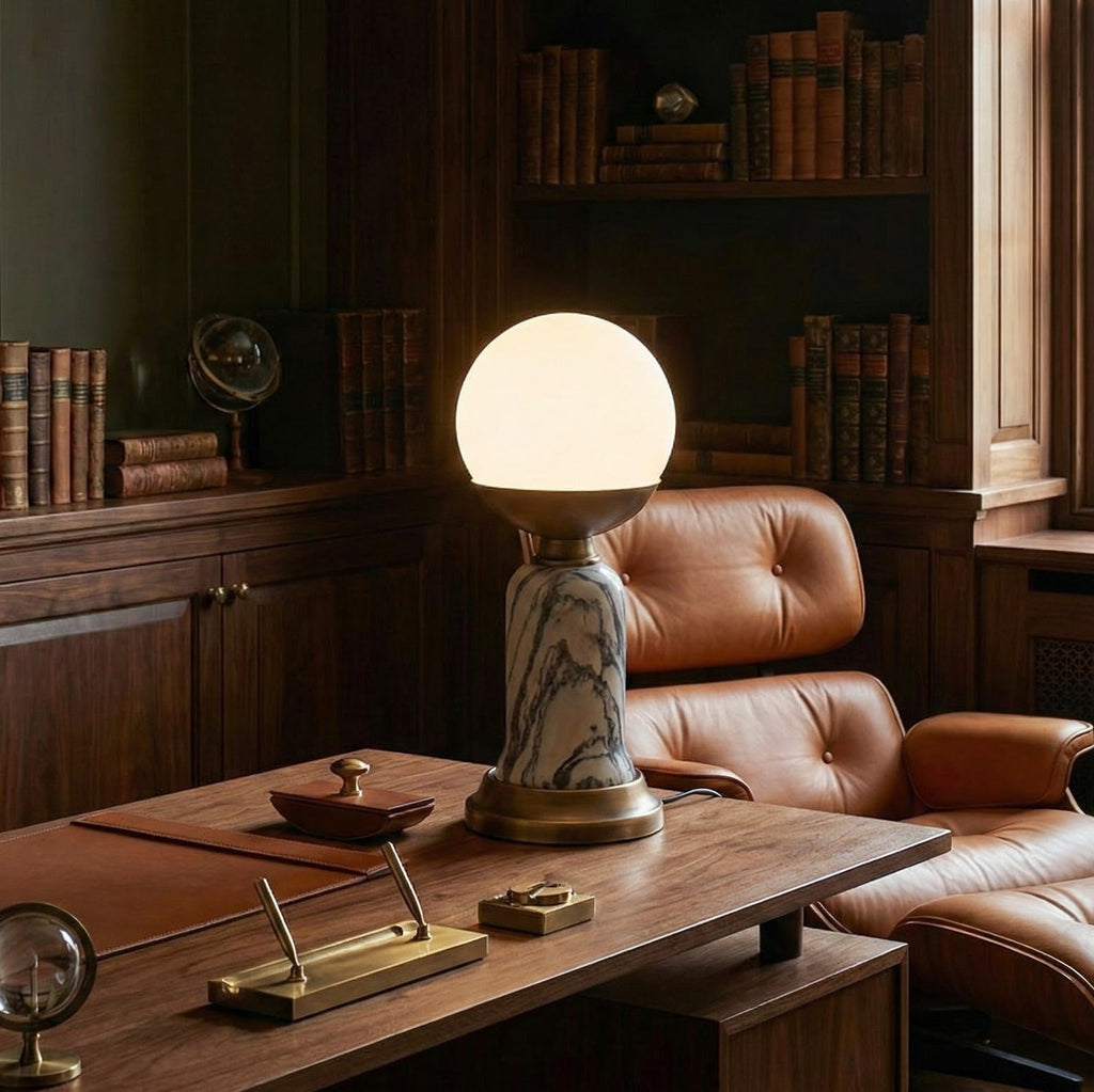 A lit Clérachic table lamp with a white globe shade on a walnut desk, beside a tan leather armchair and a wall of books.