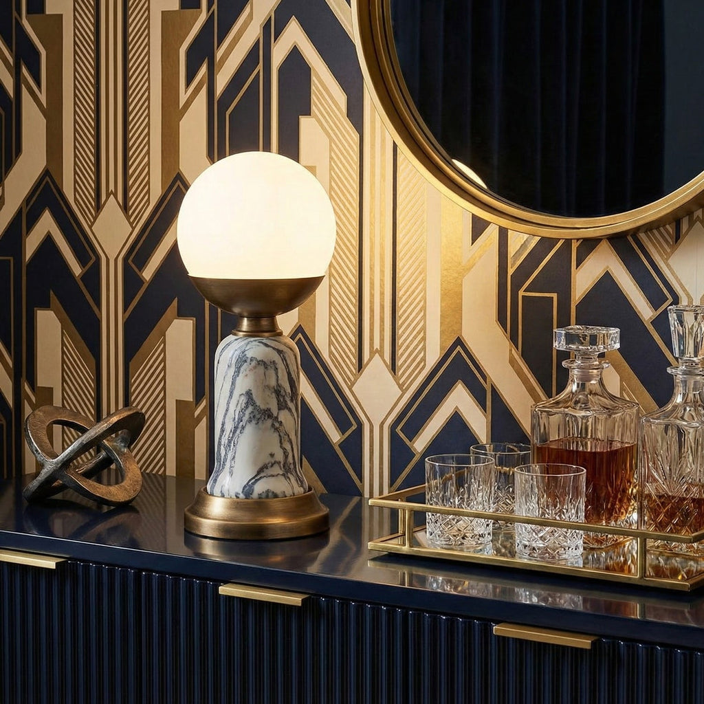On a navy cabinet beside a crystal decanter tray, a Clérachic marble-stem table lamp glows against a gold geometric wallpaper.