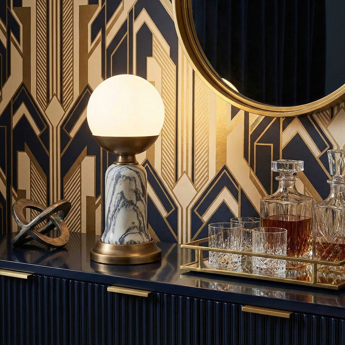 On a navy cabinet beside a crystal decanter tray, a Clérachic marble-stem table lamp glows against a gold geometric wallpaper.