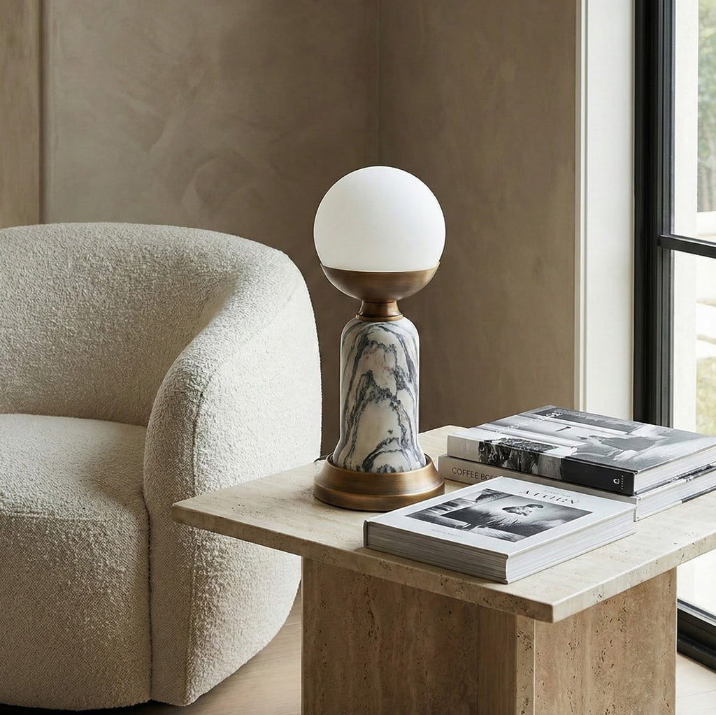 A Clérachic table lamp with a white globe on a travertine side table, next to a stack of books and a cream bouclé armchair.