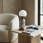 A Clérachic table lamp with a white globe on a travertine side table, next to a stack of books and a cream bouclé armchair.
