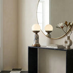 A Clérachic table lamp on a black console beneath a round mirror, with a sculptural white vase and a magazine alongside.