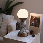 Shown here, a lit Clérachic table lamp on a travertine block, with a stone card holder, pine cones and a bouclé cushion behind it.