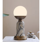 Close to the camera, a lit Clérachic table lamp shows its white globe, bronze cup and veined marble stem on a pale stone surface.
