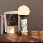 In this photo, a lit Clérachic table lamp on a wooden shelf, with a framed fashion print and a stone card holder behind it.