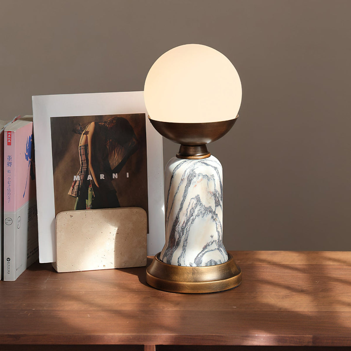 In this photo, a lit Clérachic table lamp on a wooden shelf, with a framed fashion print and a stone card holder behind it.