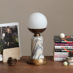 An unlit Clérachic table lamp with an opaque white globe on a wooden shelf, with a magazine, pine cones and a stack of books.