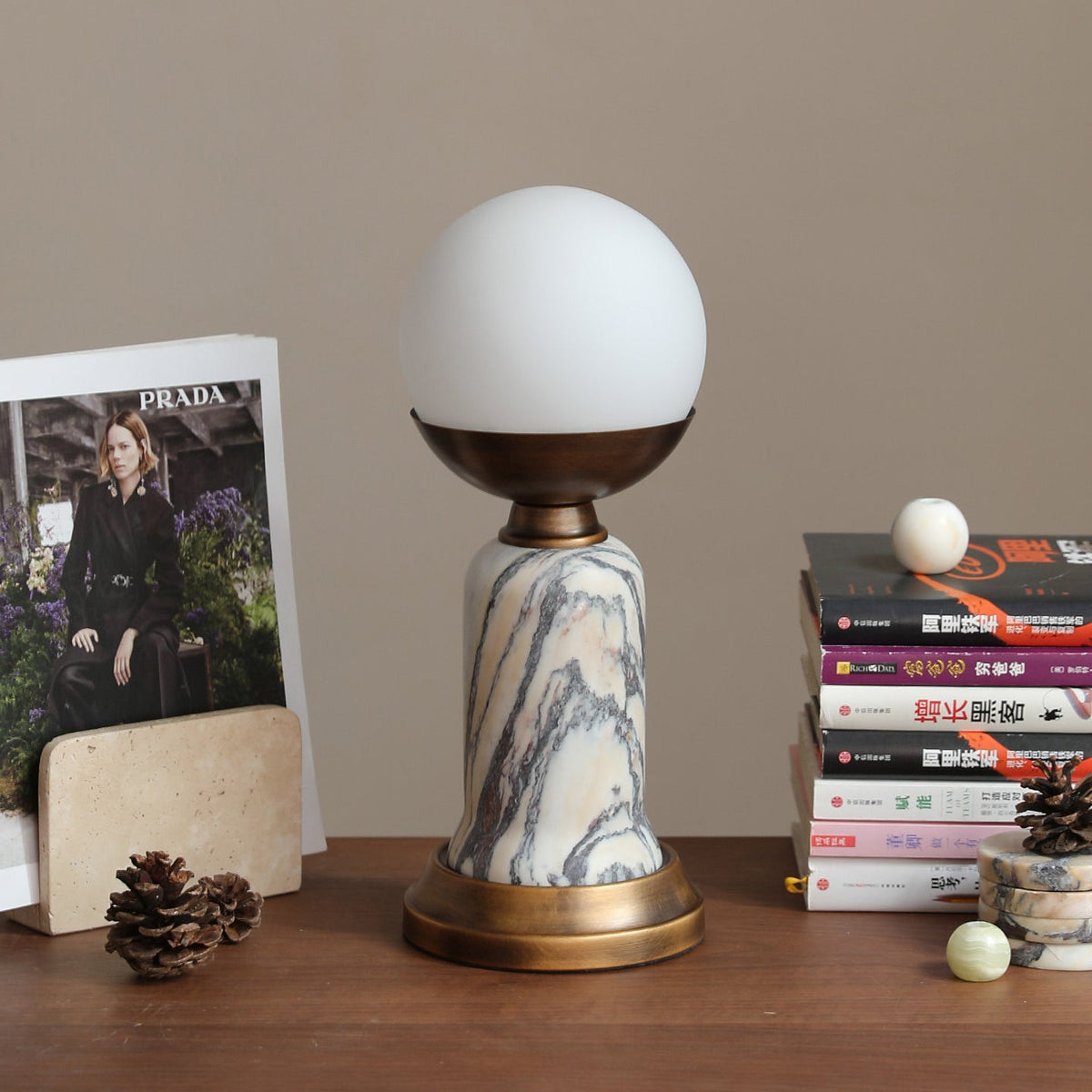 An unlit Clérachic table lamp with an opaque white globe on a wooden shelf, with a magazine, pine cones and a stack of books.