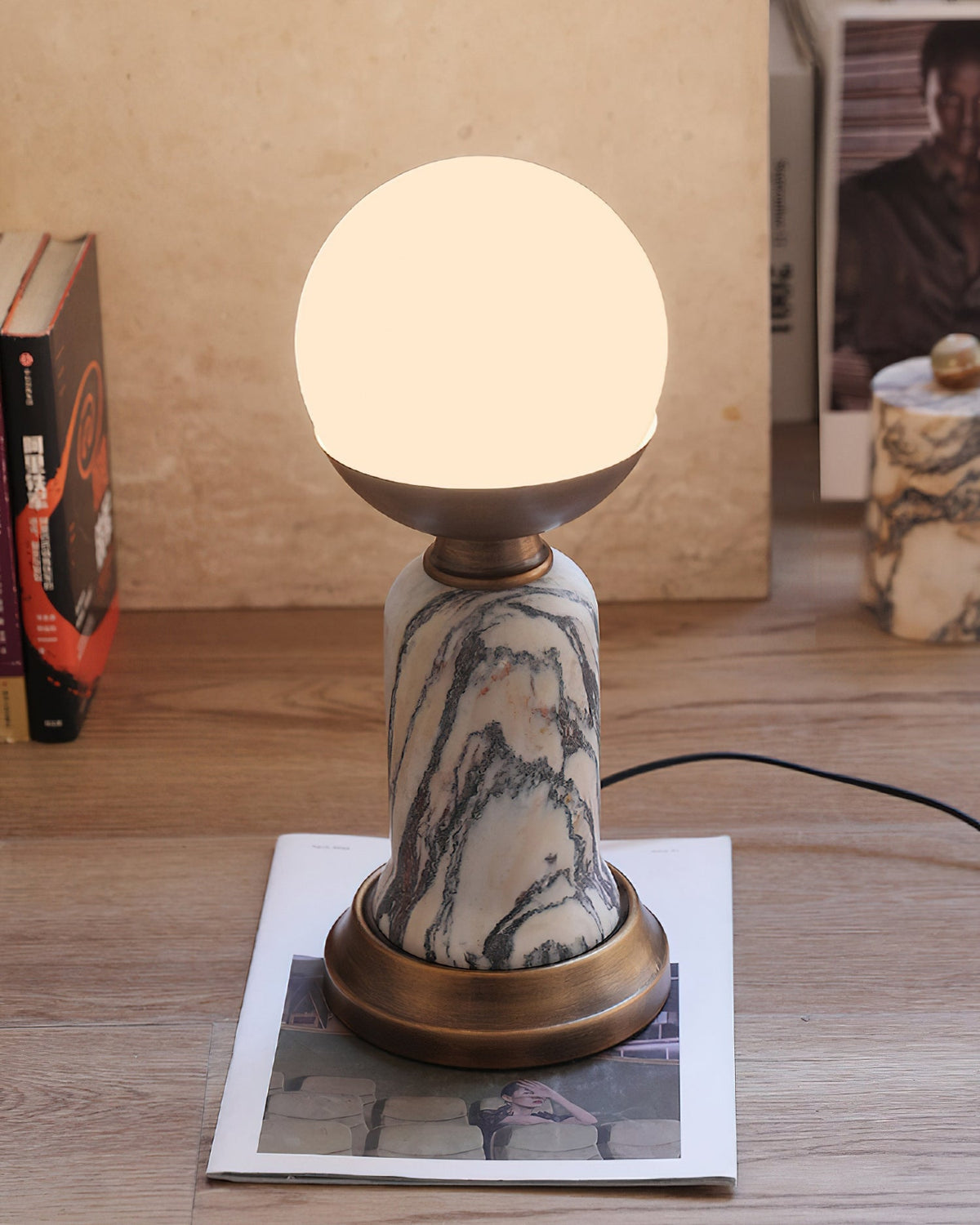 Pictured here, a lit Clérachic table lamp on a wood floor, with its black cord trailing to the right and a magazine under its base.