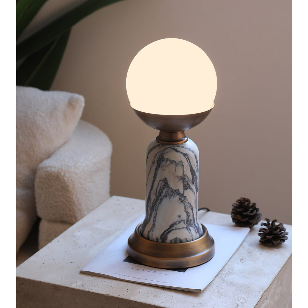 A lit Clérachic table lamp on a travertine surface, with two pine cones, a sheet of paper and a bouclé cushion behind.
