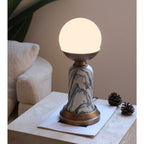 A lit Clérachic table lamp on a travertine surface, with two pine cones, a sheet of paper and a bouclé cushion behind.
