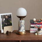 Side view of an unlit Clérachic table lamp on a wooden shelf, with a magazine, a stack of books and two small marble spheres.