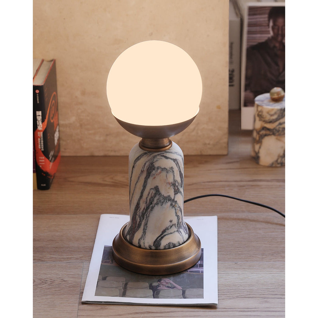 In this photo, a lit Clérachic table lamp on a wooden floor, with books and a small marble pot in the background and a cord on the right.