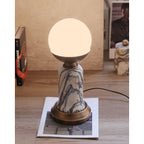 In this photo, a lit Clérachic table lamp on a wooden floor, with books and a small marble pot in the background and a cord on the right.