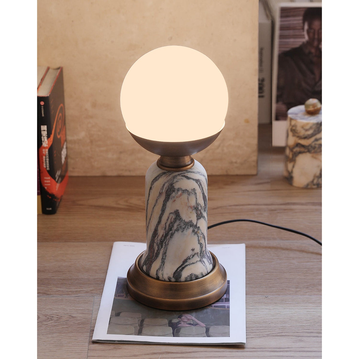In this photo, a lit Clérachic table lamp on a wooden floor, with books and a small marble pot in the background and a cord on the right.