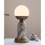 Pictured here, a lit Clérachic table lamp on a pale stone surface, with a small marble pot and a marble ball beside its bronze base.