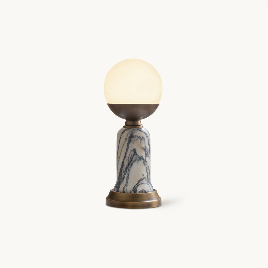 Shown here, a lit Clérachic table lamp with a glowing white globe, a bronze cup, a marble stem and a bronze base, on a cream backdrop.