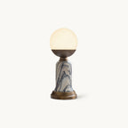 Shown here, a lit Clérachic table lamp with a glowing white globe, a bronze cup, a marble stem and a bronze base, on a cream backdrop.