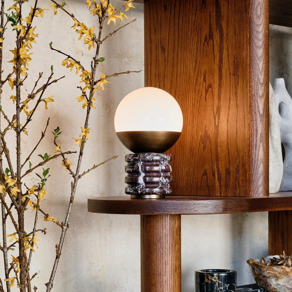 A small Clérachic globe lamp lit on a wooden shelf, beside a branch of yellow blossom and a dark oak cabinet.