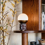 A small Clérachic globe lamp lit on a wooden shelf, beside a branch of yellow blossom and a dark oak cabinet.