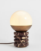 Clérachic table lamp lit on a light backdrop, its warm globe above a bronze cap, with a brown cord running to the left.