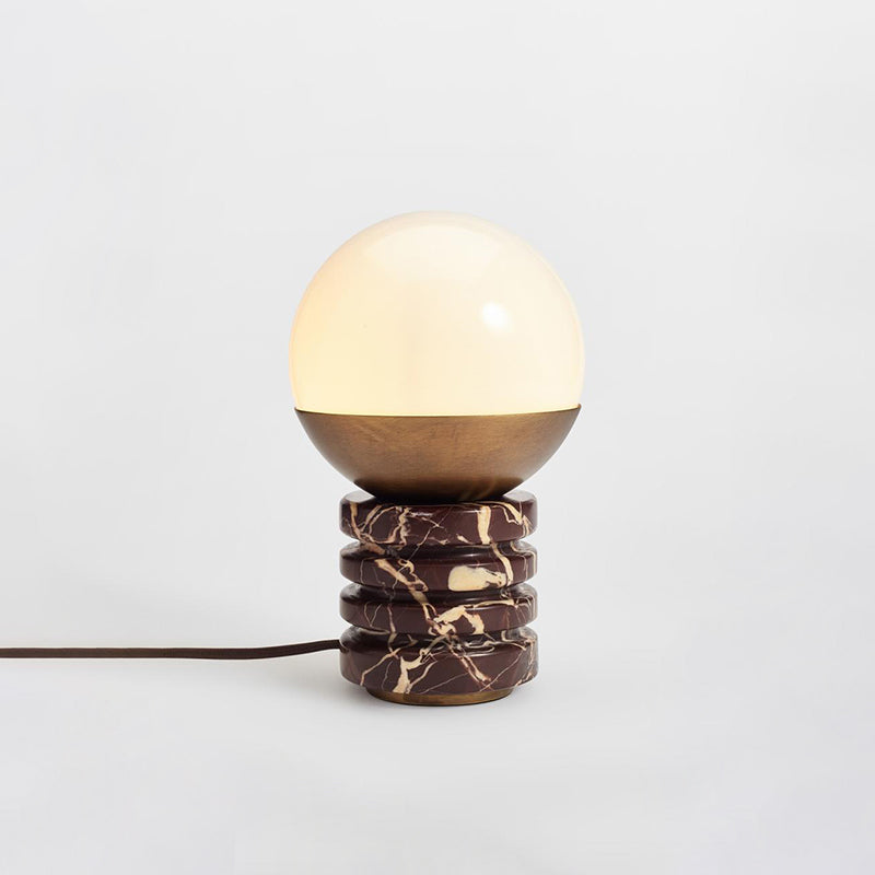Distant view of this Clérachic table lamp lit on a pale grey backdrop, its brown cord trailing out of the frame at the left.
