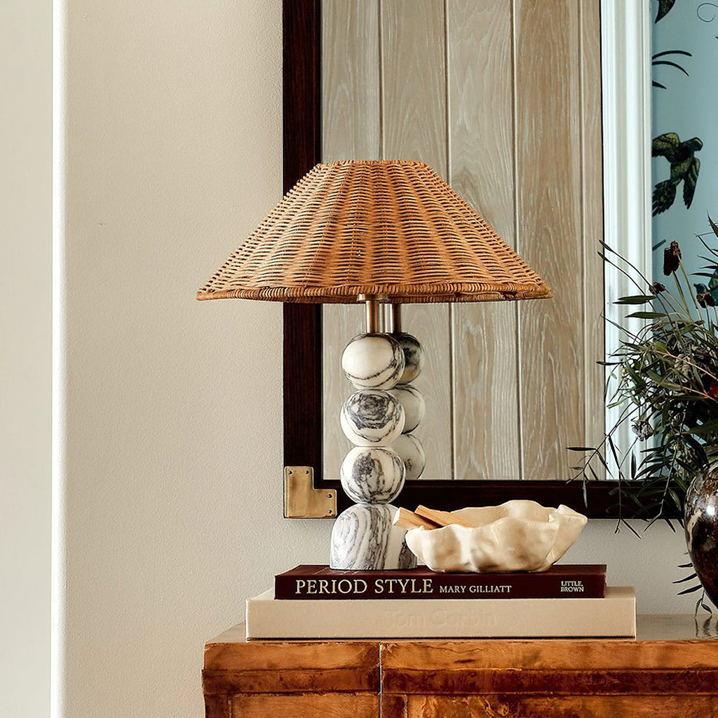 Clérachic table lamp with a stacked marble base and rattan shade on a burr wood cabinet, in front of a dark-framed mirror.