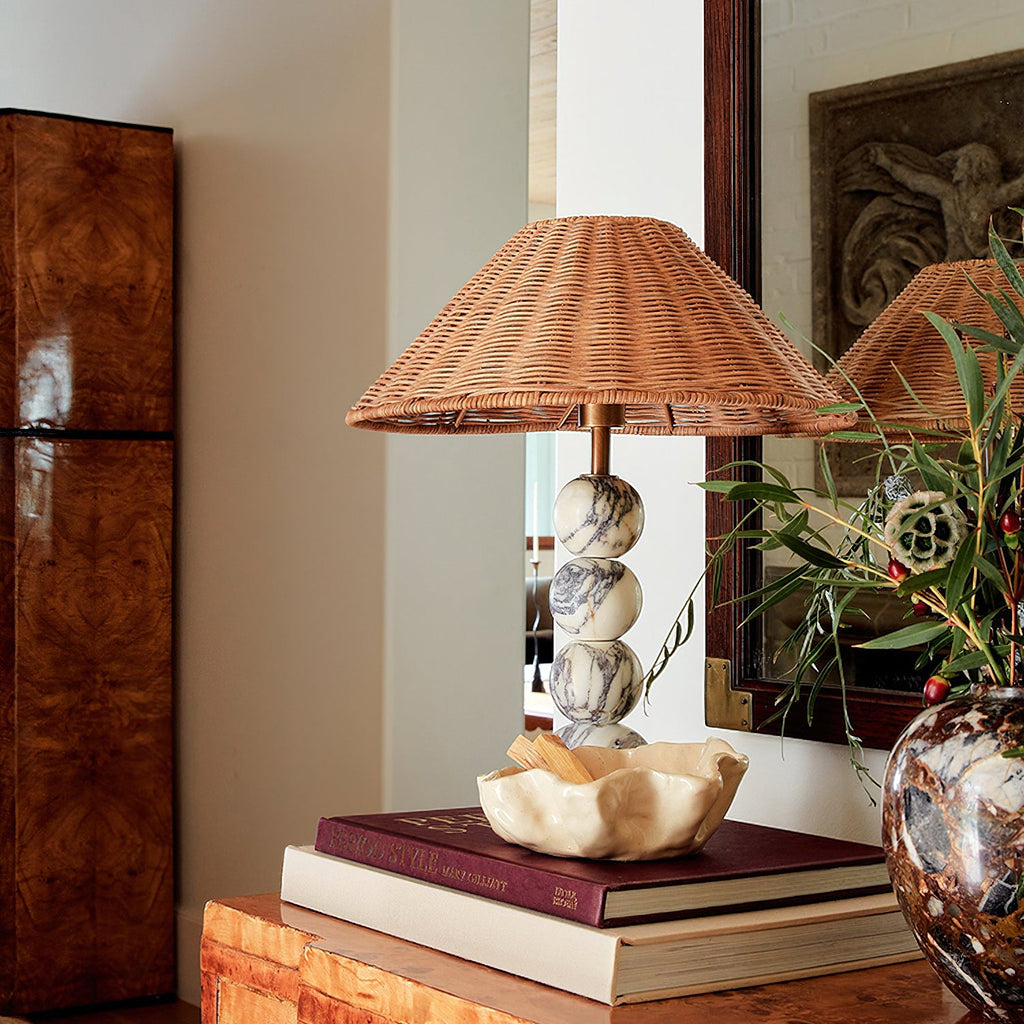 Rattan-shaded Clérachic lamp with marble spheres on a stack of books beside a cream bowl, with a walnut cabinet behind.