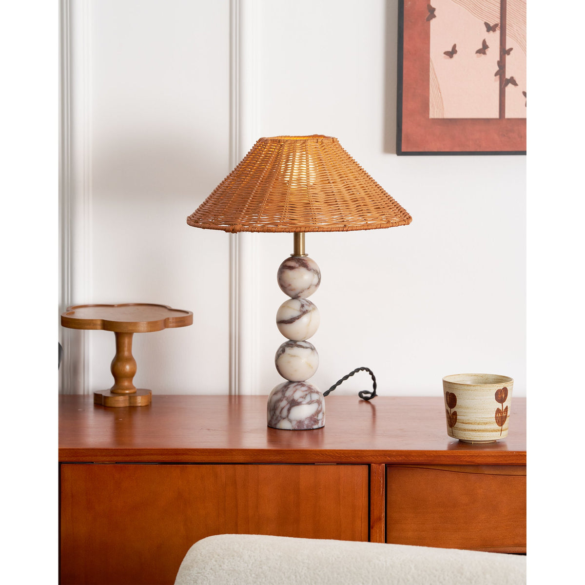 Clérachic rattan table lamp with a veined marble base on a wood sideboard, a twisted black cord and a painted cup beside it.
