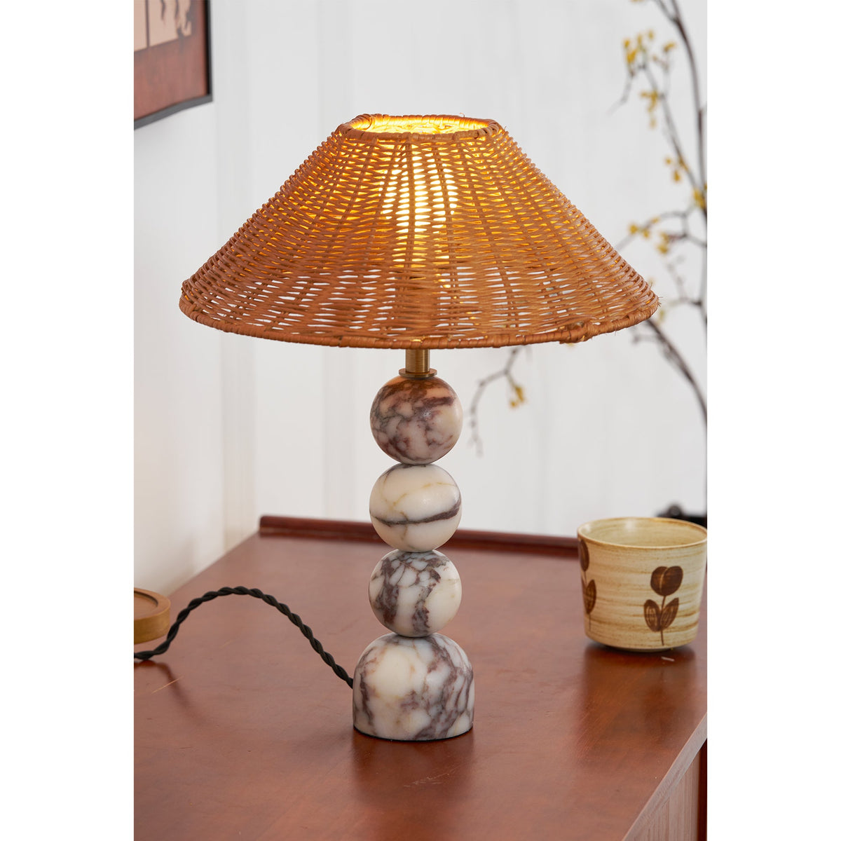 Close view of a lit Clérachic lamp on a teak sideboard, its marble spheres beside a hand-painted cup and a twisted cord.