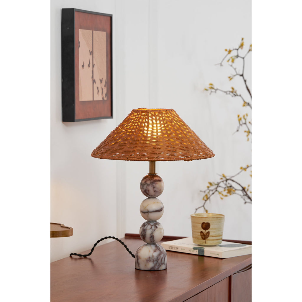 Portrait view of a lit Clérachic rattan lamp on a wooden sideboard, under a framed butterfly print and a blossom branch.