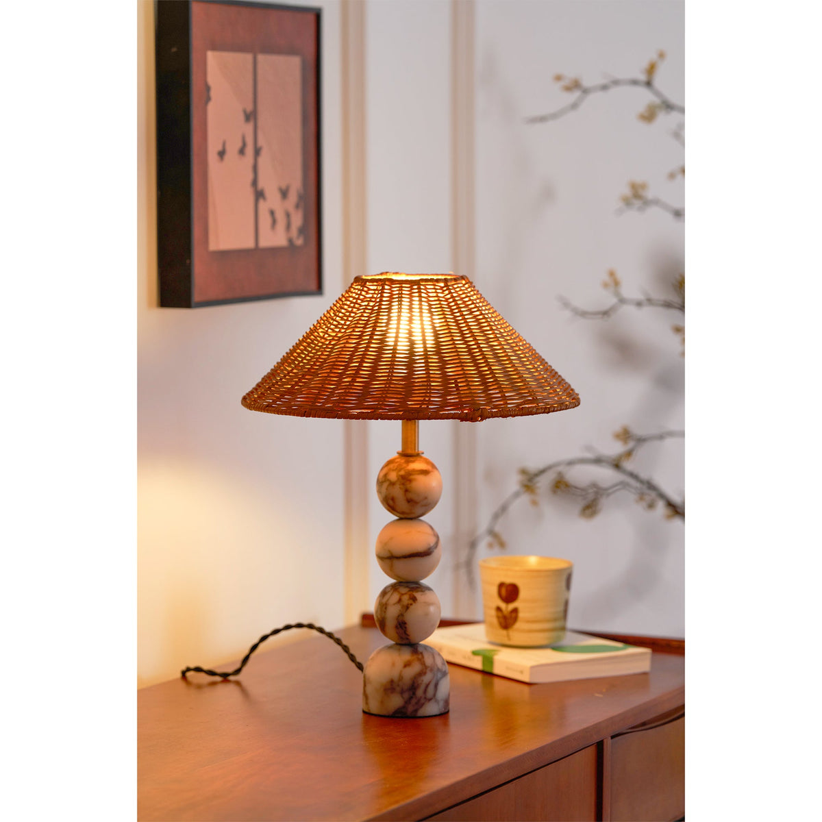 Clérachic table lamp lit above a teak sideboard, with a framed butterfly print behind and a cup resting on a book.