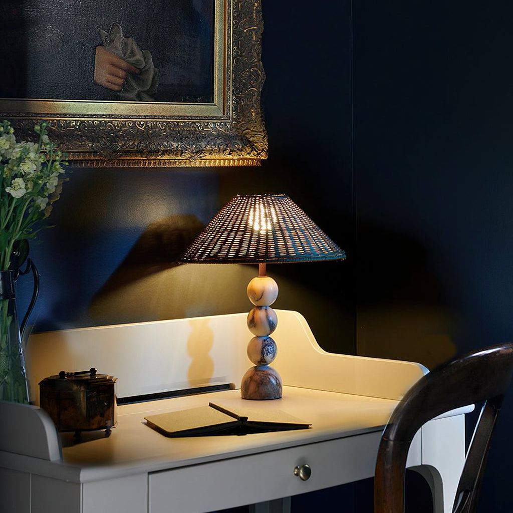 Clérachic lamp with a rattan shade on a small white desk against a deep navy wall, with an open notebook and flowers.