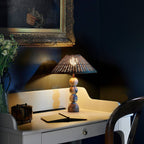 Clérachic lamp with a rattan shade on a small white desk against a deep navy wall, with an open notebook and flowers.