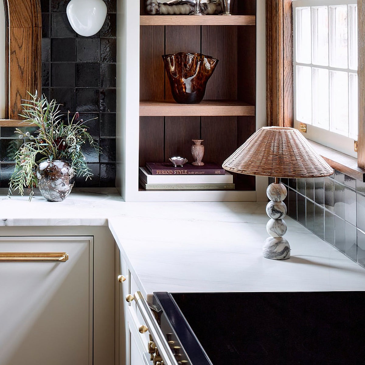 Clérachic rattan table lamp on a white stone kitchen worktop, with open shelves, a glass vase and a plant behind it.