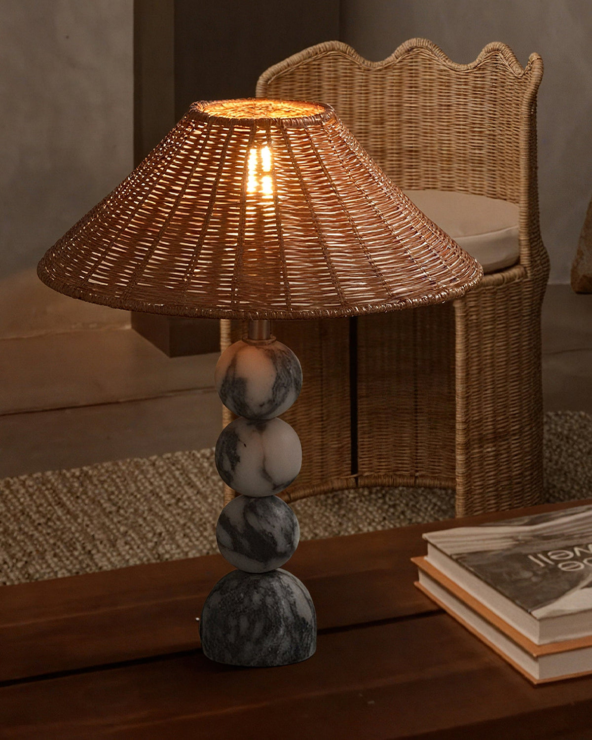 Lit Clérachic table lamp with a darker marble base on a wooden table, in front of a wicker chair and a stack of books.