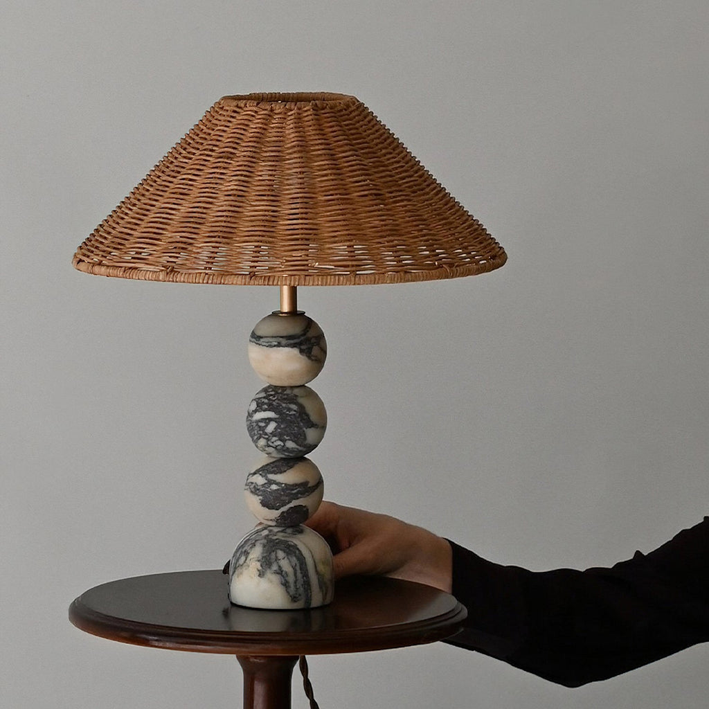 A hand resting on the marble base of this Clérachic table lamp, which stands on a small round wooden side table.