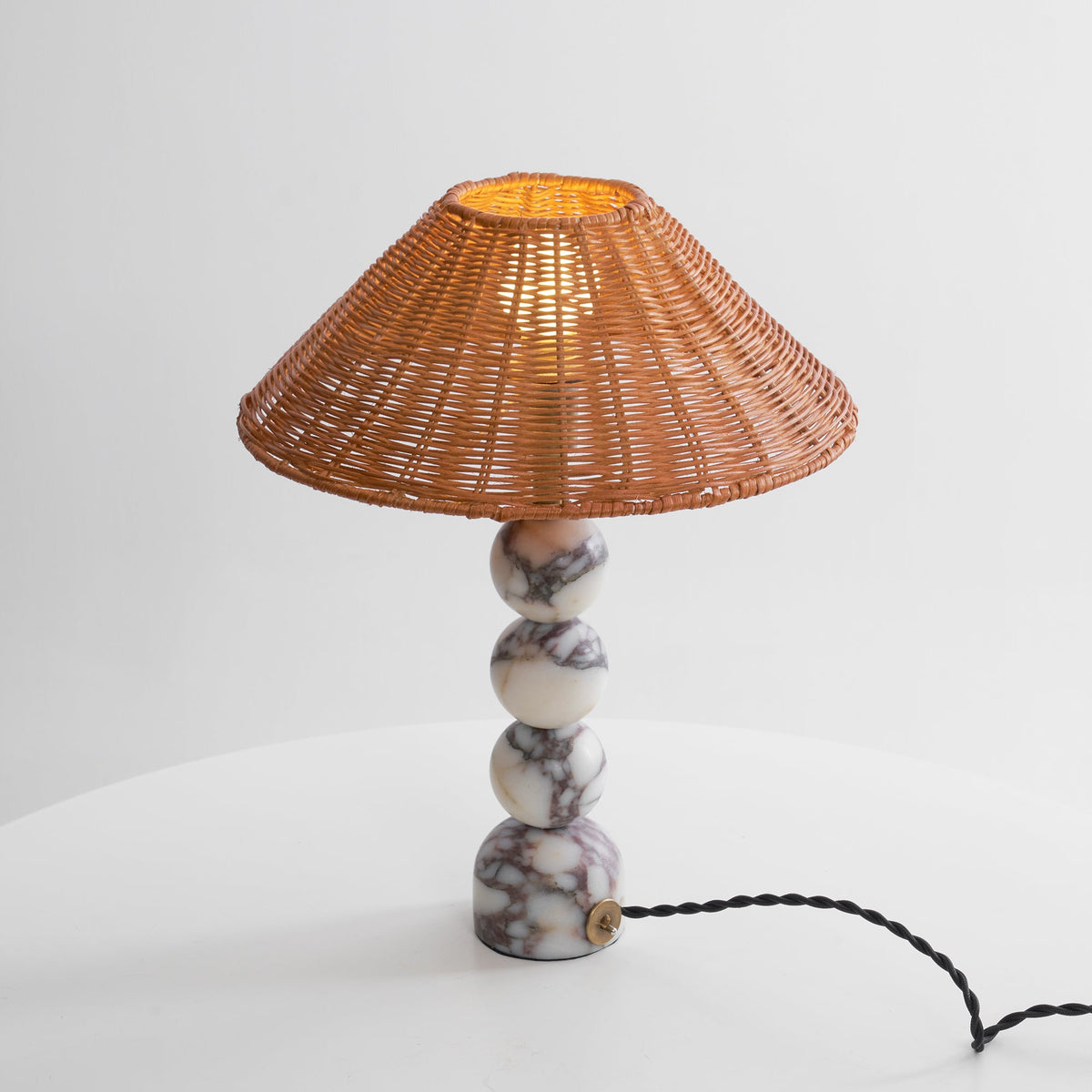 Clérachic table lamp lit from within on a white surface, its rattan shade glowing above veined marble spheres.