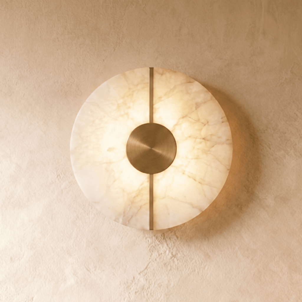 A round Clérachic alabaster wall sconce glows warmly on a plastered wall, its brushed brass disc and bar centered.