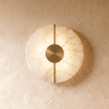 A round Clérachic alabaster wall sconce glows warmly on a plastered wall, its brushed brass disc and bar centered.
