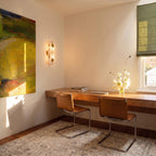A Clérachic alabaster sconce lights a sunlit home office corner beside a floating walnut desk and leather chairs.
